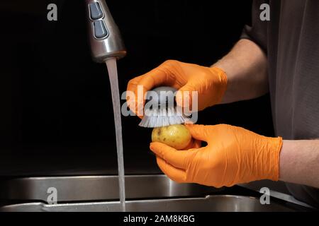 Gros plan sur le lavage et le rinçage des pommes de terre en préparation à la cuisson. Modèle avec gants en caoutchouc orange jetables. Banque D'Images