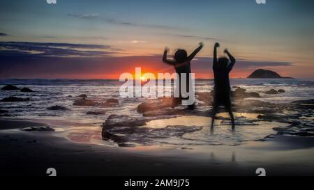 Les enfants sautant sur la plage de Muriwai au coucher du soleil, à l'ouest d'Auckland, en Nouvelle-Zélande Banque D'Images