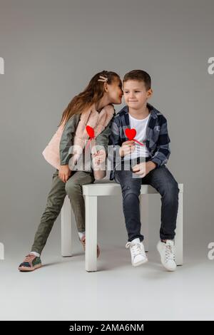 Jolie petite fille et garçon élégante avec des coeurs rouges sur le bâton dans des vêtements à la mode sitting ensemble au studio. Petite fille chople garçon et lui dit Banque D'Images