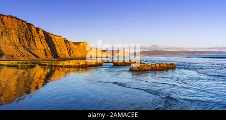 Vue sur le coucher du soleil sur le littoral de l'océan Pacifique, avec des falaises de couleur dorée reflétées sur le sable humide, Drakes Beach, point Reyes National Seashore, Califor Banque D'Images