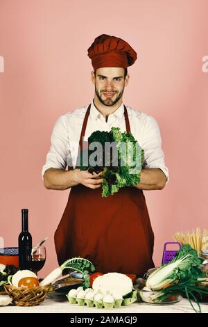 Homme en chapeau de cuisinier et tablier avec salade. La cuisine est équipée de légumes et d'outils. Ustensiles de cuisine et concept de cuisine. Le chef au visage heureux tient des petits pains de laitue et de persil sur fond rose. Banque D'Images