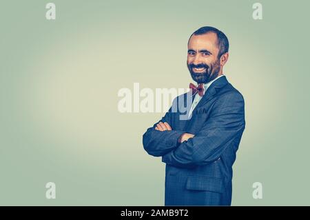 Homme souriant, confiant et barbu debout en profil avec des bras croisés. Homme d'affaires avec costume sombre, chemise bleue, noeud papillon rouge, regardant à vous appareil photo sur g Banque D'Images