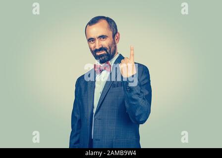 Homme d'affaires moderne et barbu montrant avec index numéro un. Gestes humains. Homme confiant et debout avec costume sombre, chemise bleue, noeud papillon rouge. Uni Banque D'Images