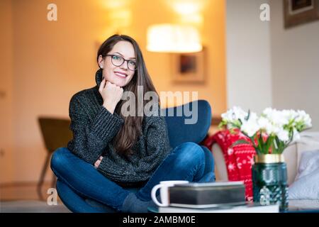 Portrait photo de la jeune femme souriante brunette se reposant sur la chaise dans le salon à la maison. Banque D'Images