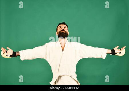 Maître de combat tient les mains larges et regarde en gratitude. Concept de victoire. Karate homme avec un visage béni dans des gants de boxe uniformes et dorés. Homme avec barbe dans kimono blanc sur fond vert. Banque D'Images