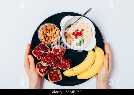 Vue de dessus femme mains tenant plaque avec petit déjeuner sain de flocons d'avoine avec amandes, bananes et graines de grenade sur table en bois blanc. Banque D'Images