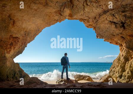 homme avec sac à dos debout dans une grotte donnant sur la mer, randonnée aventure. Banque D'Images