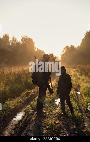Chasseurs avec équipement de chasse allant à travers le champ rural vers la forêt au coucher du soleil pendant la saison de chasse dans la campagne Banque D'Images