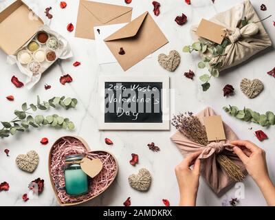 Concept de la Saint-Valentin zéro déchet. Tissu cadeau écologique enveloppant dans le style Furoshiki, bonbons faits maison et bougie comme idées cadeaux et tableau de surveillance avec lettres de Saint-Valentin zéro déchet sur table en marbre Banque D'Images