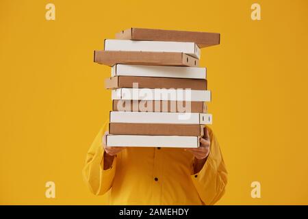 Taille vers le haut portrait de livreur inconnu se cachant derrière des boîtes à pizza tout en se tenant sur fond jaune pop dans le studio Banque D'Images