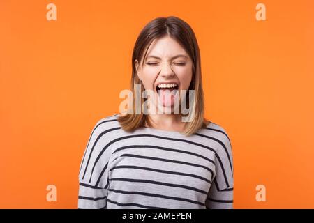 Portrait d'une femme amusante et insouciante avec des cheveux bruns dans une chemise rayée à manches longues, debout avec des yeux fermés, montrant sa langue et son grimace. IND Banque D'Images