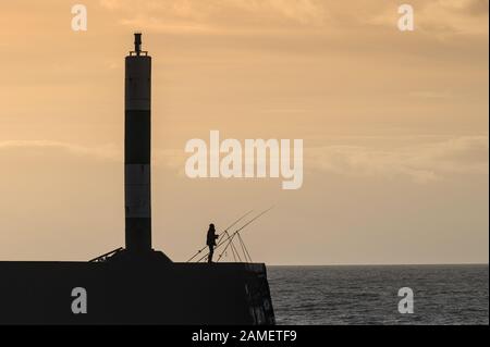 Pêcheur de Rod and Line en silhouette sur la jetée en pierre d'Aberystwyth sur un fond de ciel orange Banque D'Images