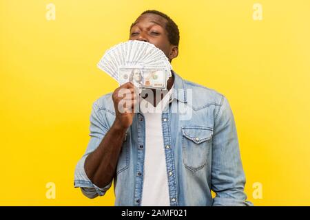 Portrait de jeune homme avide dans une chemise en denim debout avec des yeux fermés et des factures de dollar odorant avec une expression satisfaite heureuse, cachant demi-face. I Banque D'Images