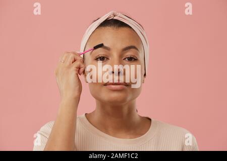 Tête et épaules portrait de belle femme mixte de course se brossant les sourcils tout en mettant le maquillage le matin debout contre fond rose poussière, espace de copie Banque D'Images