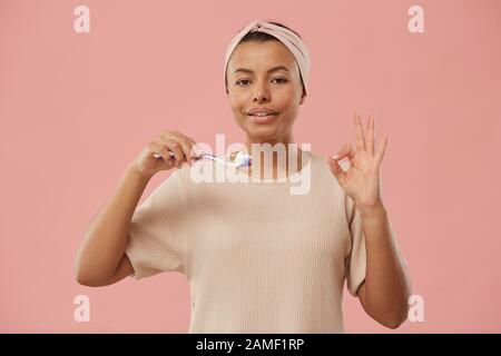 Taille vers le haut portrait de belle femme de course mixte tenant la brosse à dents et montrant OK signe le matin tout en se tenant contre poussière rose fond, espace de copie Banque D'Images