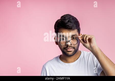 Jeune homme indien sérieux avec mauvaise vue, regardant soigneusement à travers les lunettes, essayant de lire quelque chose, touchant le cadre de lunettes, portant décontracté ou Banque D'Images