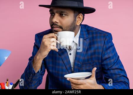 Gros plan de jeune homme d'affaires indien non rasé dans un costume à damier bleu formel, chapeau noir , assis sur le lieu de travail, pensant à quelque chose, Banque D'Images
