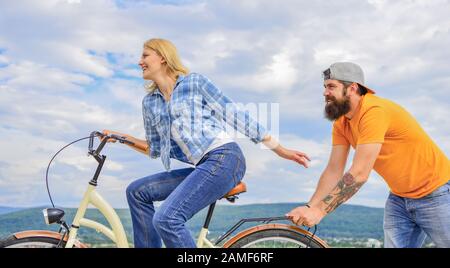 Vélo fille tandis que l'homme la soutenir. Aide à croire en vous-même. Impulsion L'impression de commencer à bouger. Des promenades en vélo femme fond de ciel. Pousser et la promotion. Impulsion à se déplacer. Homme pousse fille faire du vélo. Banque D'Images