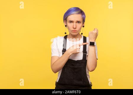 Portrait d'une fille hipster mécontente en colère avec des cheveux violets dans des salopettes en denim pointant sur son poignet et regardant impatiente, ce qui signifie pas de temps, aussi Banque D'Images