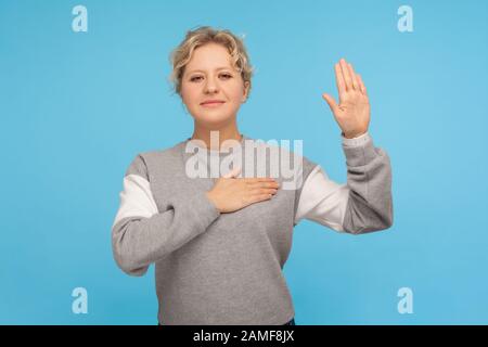 Portrait de citoyen patriotique, honnête femme adulte avec des cheveux bouclés dans le sweat-shirt en gardant la main sur la poitrine et en levant la paume, en faisant la promesse de fidélité, serment. Banque D'Images