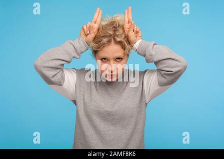 Femme agressive en sweat-shirt gris montrant des cornes de taureau gestes avec les doigts sur la tête et de puffer les joues, menaçant d'attaquer, exprimant la colère. Banque D'Images