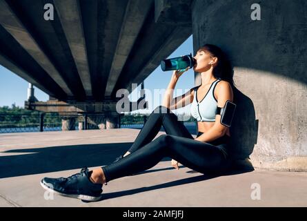 Jeune femme de fitness buvant de l'eau de la bouteille. Une femme sportive se reposant après une activité physique dans une ville industrielle à proximité de la rivière Banque D'Images