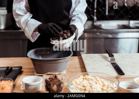 vue rognée du chocolat dans des gants en latex ajoutant des copeaux de chocolat noir dans un bol sur une balance de cuisine Banque D'Images