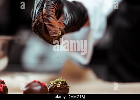 vue réduite du chocolat dans un gant en latex noir ajoutant de la poudre de pistache verte sur des bonbons frais Banque D'Images