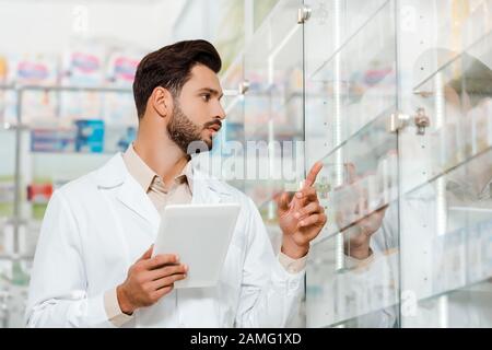 Un beau pharmacien avec tablette numérique pointant vers la vitrine en pharmacie Banque D'Images