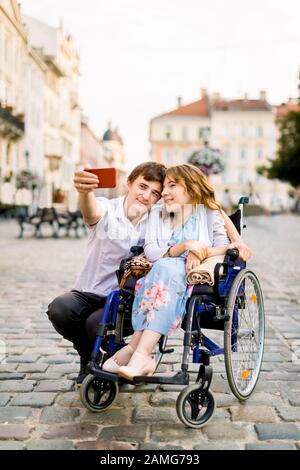 Jeune femme en fauteuil roulant et son mari prenant selfie dehors, marchant dans le vieux centre-ville Banque D'Images