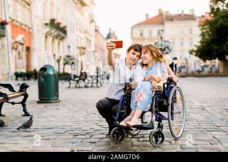 Heure du selfie. Beau jeune homme heureux embrassant sa jeune femme handicapée et souriant tout en prenant selfie avec elle dans la ville Banque D'Images