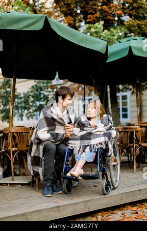 Deux amis, un couple charmant homme et une femme couverts de carreaux dans un café en plein air buvant du café, l'homme assis en fauteuil roulant Banque D'Images