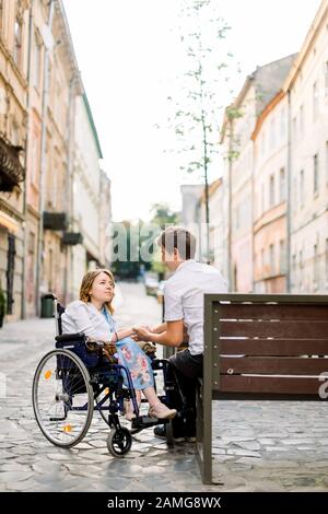 bel homme et jolie femme en fauteuil roulant se regardant l'un l'autre dans la rue. Un couple charmant dans les cheveux de petit-lait marchant ensemble dans la ville Banque D'Images