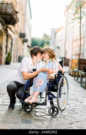 Jeune homme hople sa jolie femme sur le fauteuil roulant, tout en marchant ensemble dans la rue de la ville. Couple amoureux du fauteuil roulant embrassant et touchant Banque D'Images