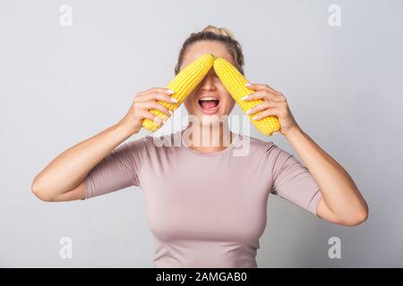 Portrait d'une femme gaie couvrant les yeux avec des épis de maïs, ouverture de la bouche surprise, concept de saine alimentation et de régime végétarien, légumes frais crus Banque D'Images