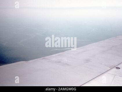 Photographie vernaculaire prise sur une transparence de film analogique de 35 mm, considérée comme représentant des nuages blancs au-dessus de la mer, avec aile d'avion commercial au premier plan, 1965. Les principaux sujets/objets détectés sont le ciel, le paysage, l'hiver, la nature et la couleur grise. () Banque D'Images