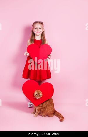 Petit enfant heureux en robe rouge et chat britannique rouge avec un coeur en papier sur fond rose Banque D'Images