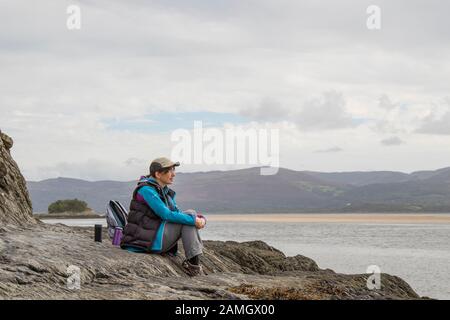 Jeune femme isolée en casquette de marche et sport, assise sur des rochers au bord de la côte, vue sur la mer, profondément dans la pensée, pays de Galles Royaume-Uni. Recharge des batteries. Banque D'Images