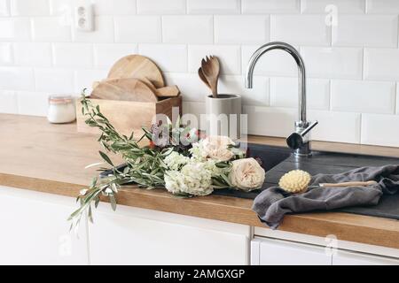 Gros plan de l'intérieur de la cuisine. Mur de briques blanches, carrelage de métro, comptoirs en bois avec ustensiles de cuisine. Roses fleurs dans évier noir. scandinave moderne Banque D'Images