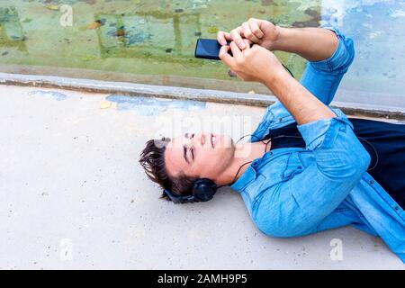 Jeune gars dans un casque noir couché sur le sol rêveusement en utilisant le téléphone portable passer du temps au parc Banque D'Images