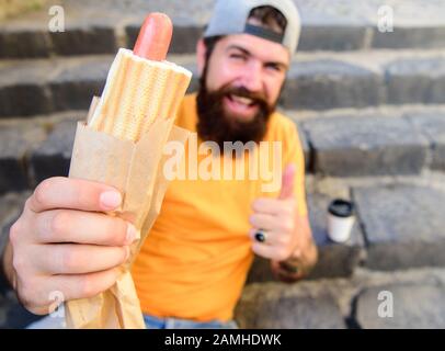 Homme barbu profiter de la nourriture de rue escalier arrière-plan. Hipster mange un chien chaud et boit du café. Concept de l'heure du déjeuner. Hipster appréciez la boisson chaude de votre chien et de votre tasse en papier. Restauration rapide pour déjeuner idée commune. Banque D'Images