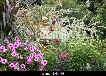 Statue peinte en or de jeune garçon jouant une mandoline à la frontière avec des fleurs roses Petunia x hybrida 'Easy Wave Plum Vein', rouge Rosa - Roses et Salix. Banque D'Images