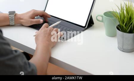 Photo courte d'un freelance masculin en tapant sur une tablette dans un espace de travail simple avec une tasse à café et une cafetière sur une table en bois blanc Banque D'Images