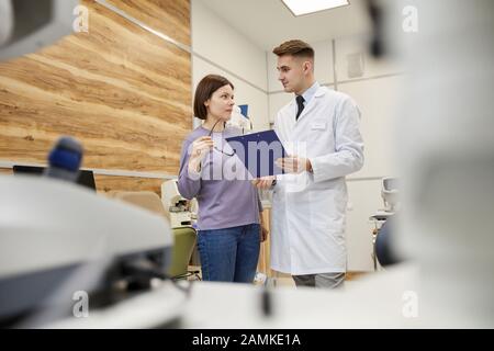 Portrait de beau jeune ophtalmologiste tenant le presse-papiers tout en consultant patient féminin dans la clinique moderne, espace de copie Banque D'Images