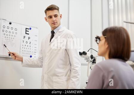 Portrait d'un jeune optométriste pointant vers un graphique de vision tout en vérifiant la vue d'une patiente dans une clinique moderne d'ophtalmologie Banque D'Images