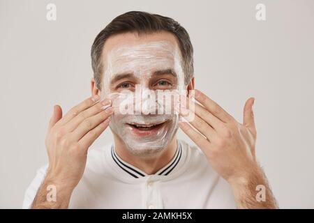 Tête et épaules portrait de l'homme adulte souriant appliquant le masque de visage tout en appréciant la routine du matin debout contre fond blanc Banque D'Images