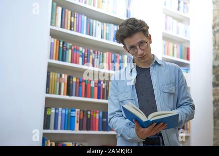 Jeune homme sérieux avec des lunettes, regardant de la lecture, à côté d'une étagère Banque D'Images