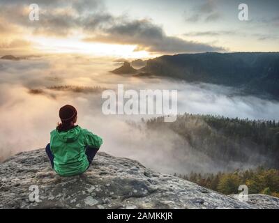 Belle femme de corps assis dans un windcheater vert sur le sommet de montagne regardant le lever du soleil sur une mer de brouillard Banque D'Images