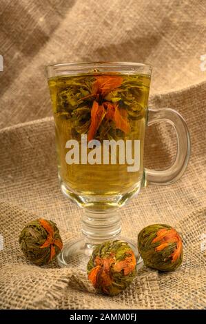 Thé fleuri préparé dans une tasse de verre et des boules de thé de fleur sur un fond de tissu homespun rugueux. Gros plan Banque D'Images