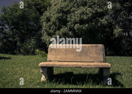 Photographie d'un banc de parc en béton sur la pelouse avec des arbres derrière. Banque D'Images